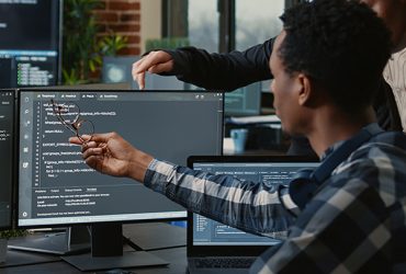 Software developers discussing about source code compiling discovers errors and asks the rest of the team for explanations in front of multiple screens running algorithms. Programmers doing teamwork.