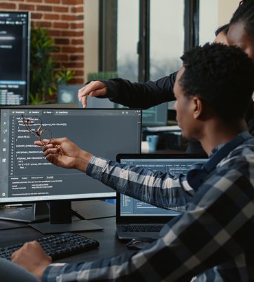 Software developers discussing about source code compiling discovers errors and asks the rest of the team for explanations in front of multiple screens running algorithms. Programmers doing teamwork.